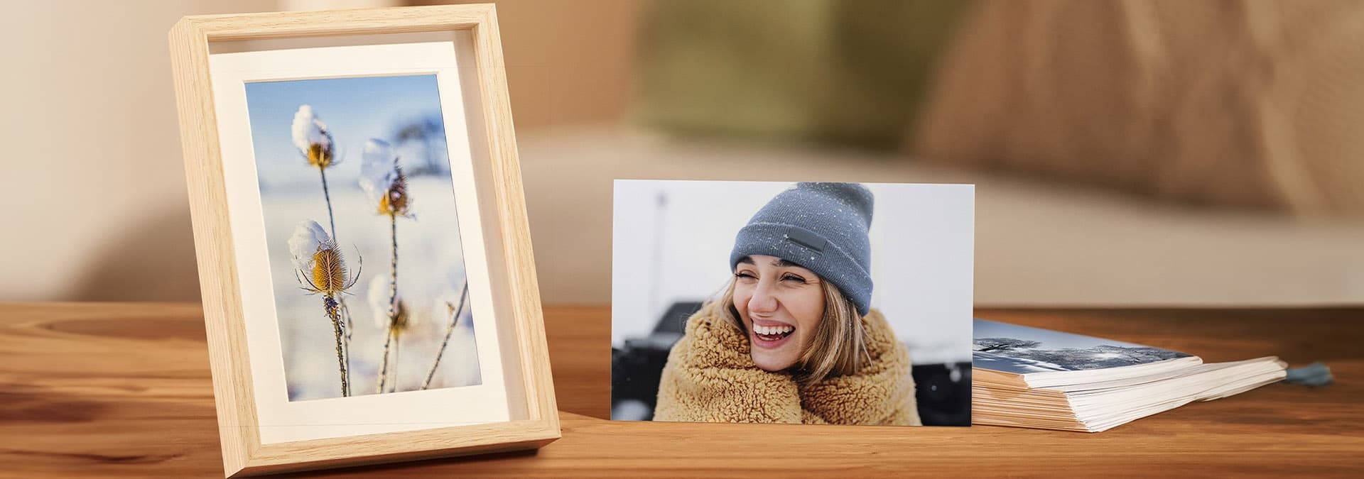 Zarámovaná zimná fotografia s detailom na bodliaky pokryté snehom vedľa fotografie osoby v zimnej čiapke a bunde na drevenom stole.