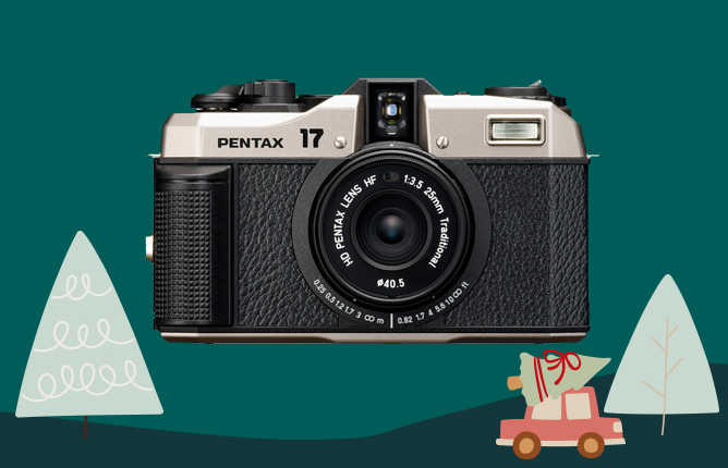 Fotoaparát Pentax 17 s čiernym koženým dizajnom, obklopený kreslenými stromčekmi a darčekom na autíčku